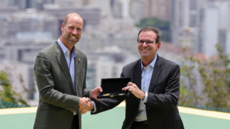 Dois homens de terno trocam um aperto de mão enquanto um deles segura uma caixa aberta com uma chave dourada em seu interior, com prédios desfocados ao fundo.