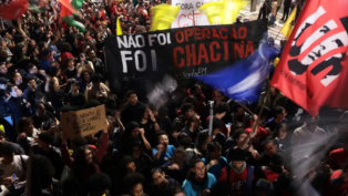 Multidão participa de manifestação à noite segurando faixas e cartazes. Um dos principais cartazes diz “Não foi operação, foi chacina”, e outro, “A juventude quer viver! Parem de nos matar!”. Bandeiras coloridas são erguidas entre os manifestantes.