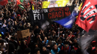Multidão participa de manifestação à noite segurando faixas e cartazes. Um dos principais cartazes diz “Não foi operação, foi chacina”, e outro, “A juventude quer viver! Parem de nos matar!”. Bandeiras coloridas são erguidas entre os manifestantes.