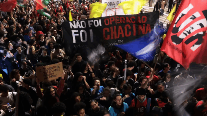 Multidão participa de manifestação à noite segurando faixas e cartazes. Um dos principais cartazes diz “Não foi operação, foi chacina”, e outro, “A juventude quer viver! Parem de nos matar!”. Bandeiras coloridas são erguidas entre os manifestantes.