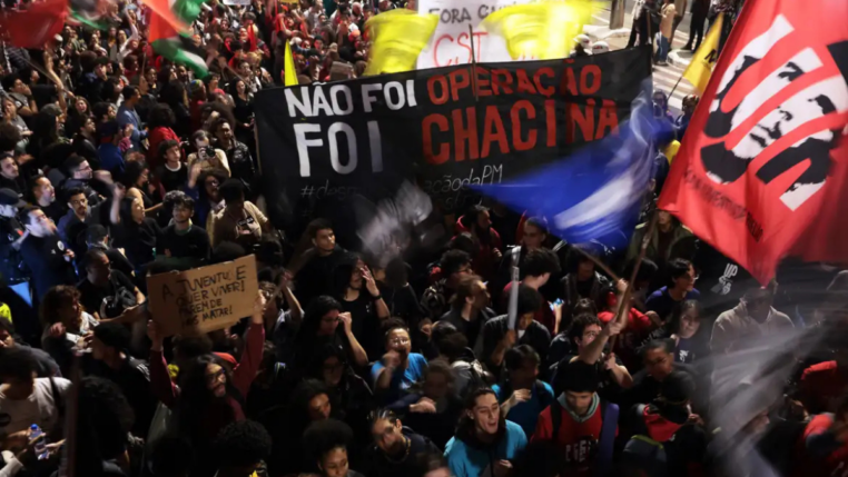 Multidão participa de manifestação à noite segurando faixas e cartazes. Um dos principais cartazes diz “Não foi operação, foi chacina”, e outro, “A juventude quer viver! Parem de nos matar!”. Bandeiras coloridas são erguidas entre os manifestantes.