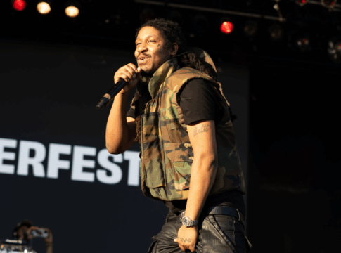 Homem segura um microfone enquanto canta em um palco durante um show ao ar livre. Ele veste colete camuflado e calça preta, com a palavra “SUMMERFEST” visível no fundo.