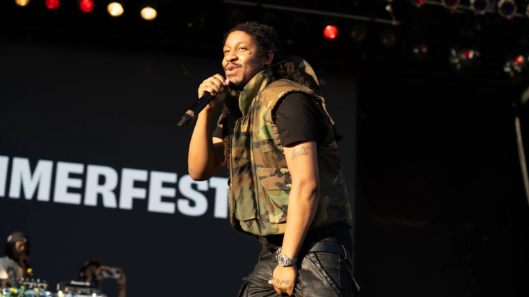Homem segura um microfone enquanto canta em um palco durante um show ao ar livre. Ele veste colete camuflado e calça preta, com a palavra “SUMMERFEST” visível no fundo.
