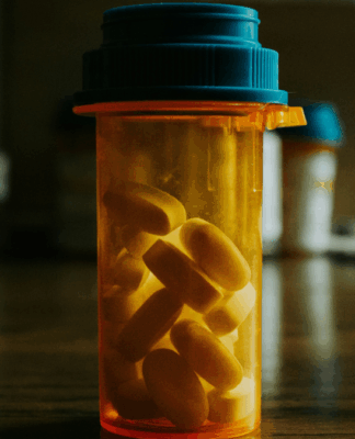 Frasco transparente com tampa azul contendo comprimidos amarelos sobre uma mesa de madeira, em ambiente interno com iluminação suave e fundo desfocado.