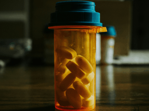 Frasco transparente com tampa azul contendo comprimidos amarelos sobre uma mesa de madeira, em ambiente interno com iluminação suave e fundo desfocado.