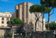 Vista diurna da Torre dei Conti em Roma, uma estrutura medieval de tijolos cercada por árvores altas e edifícios históricos sob um céu azul claro.