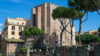 Vista diurna da Torre dei Conti em Roma, uma estrutura medieval de tijolos cercada por árvores altas e edifícios históricos sob um céu azul claro.