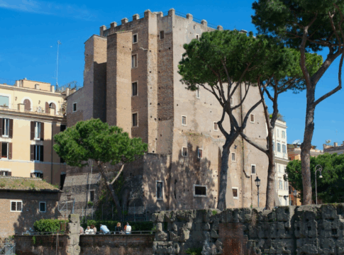 Vista diurna da Torre dei Conti em Roma, uma estrutura medieval de tijolos cercada por árvores altas e edifícios históricos sob um céu azul claro.