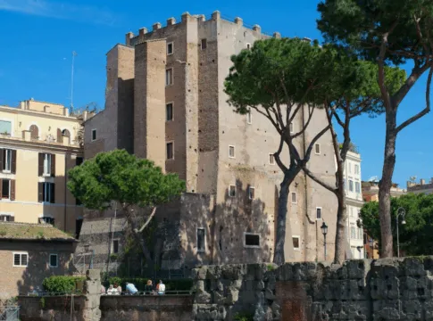 Vista diurna da Torre dei Conti em Roma, uma estrutura medieval de tijolos cercada por árvores altas e edifícios históricos sob um céu azul claro.