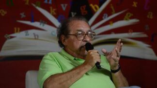 Rui Castro , vestindo camisa polo verde-clara, falando ao microfone durante um evento literário. Ao fundo, há uma imagem de um livro aberto com letras coloridas espalhadas sobre um painel vermelho