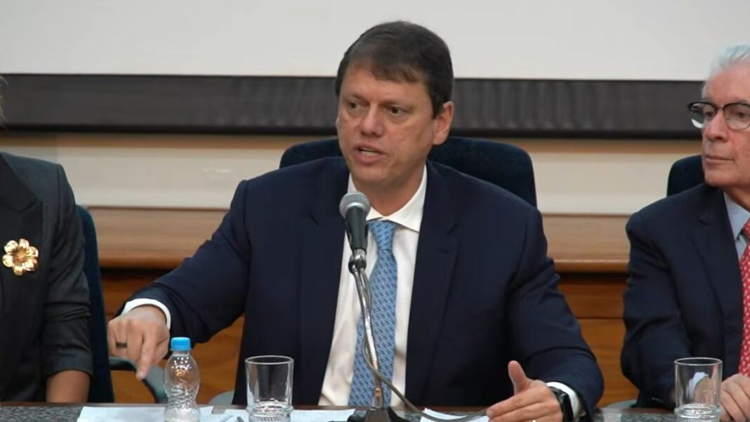 Governador Tarcísio de Freitas, de terno, sentado à mesa da coletiva e falando no microfone