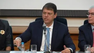 Governador Tarcísio de Freitas, de terno, sentado à mesa da coletiva e falando no microfone