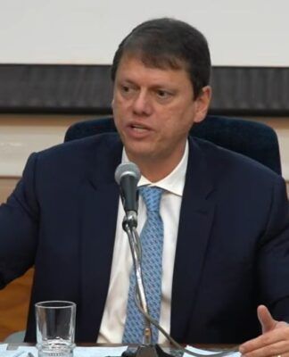 Governador Tarcísio de Freitas, de terno, sentado à mesa da coletiva e falando no microfone