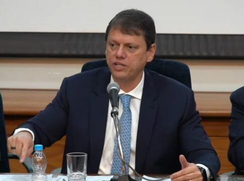 Governador Tarcísio de Freitas, de terno, sentado à mesa da coletiva e falando no microfone