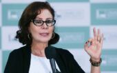 Carla Zambelli, na época em que era parlamentar, vestindo roupa formal e gesticulando com o sinal de "Ok" enquanto discursa em palanque