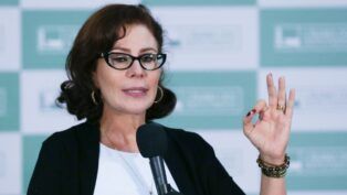 Carla Zambelli, na época em que era parlamentar, vestindo roupa formal e gesticulando com o sinal de "Ok" enquanto discursa em palanque