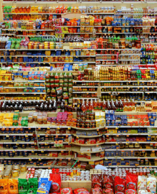 Corredor de supermercado visto de cima, com prateleiras repletas de produtos organizados por categorias e cores, formando linhas densas e simétricas de embalagens ao longo do espaço.