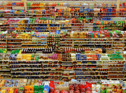 Corredor de supermercado visto de cima, com prateleiras repletas de produtos organizados por categorias e cores, formando linhas densas e simétricas de embalagens ao longo do espaço.