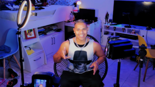 Homem sorri enquanto grava um vídeo em um quarto iluminado por luzes coloridas e equipamentos de gravação, sentado em frente a uma câmera e ring light, com prateleiras e objetos decorativos ao fundo.