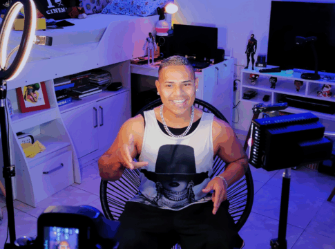 Homem sorri enquanto grava um vídeo em um quarto iluminado por luzes coloridas e equipamentos de gravação, sentado em frente a uma câmera e ring light, com prateleiras e objetos decorativos ao fundo.
