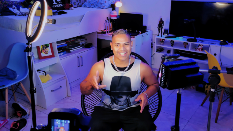 Homem sorri enquanto grava um vídeo em um quarto iluminado por luzes coloridas e equipamentos de gravação, sentado em frente a uma câmera e ring light, com prateleiras e objetos decorativos ao fundo.