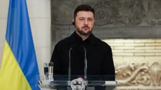 Homem vestindo roupa preta fala em um púlpito transparente equipado com microfones, usando um auricular, enquanto uma bandeira azul e amarela aparece à esquerda e um fundo de pedra esculpida compõe o cenário.