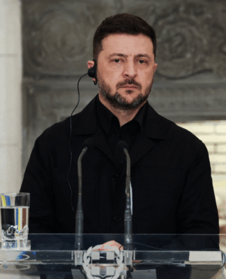 Homem vestindo roupa preta fala em um púlpito transparente equipado com microfones, usando um auricular, enquanto uma bandeira azul e amarela aparece à esquerda e um fundo de pedra esculpida compõe o cenário.