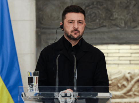 Homem vestindo roupa preta fala em um púlpito transparente equipado com microfones, usando um auricular, enquanto uma bandeira azul e amarela aparece à esquerda e um fundo de pedra esculpida compõe o cenário.