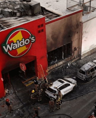 Imagem aérea mostra fachada queimada de uma loja Waldo’s, no México, após um incêndio. Bombeiros e equipes de resgate trabalham no local, ao lado de carros carbonizados e escombros espalhados pela rua.