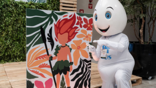 Mascote em formato de gota d’água, com olhos grandes e expressão sorridente, ajoelha-se ao lado de um painel colorido que mostra ilustração de personagem de cabelo vermelho em meio a folhas e flores tropicais, apontando para a arte.