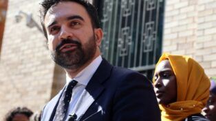 Zohran Mamdani discursando em um espaço público aberto. Atrás dele, uma mulher de jihad amarela