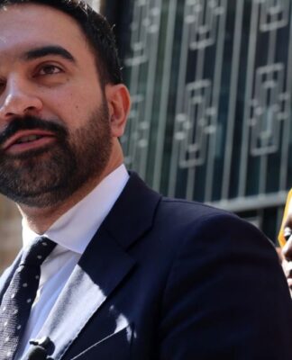 Zohran Mamdani discursando em um espaço público aberto. Atrás dele, uma mulher de jihad amarela