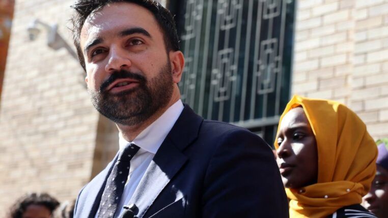 Zohran Mamdani discursando em um espaço público aberto. Atrás dele, uma mulher de jihad amarela