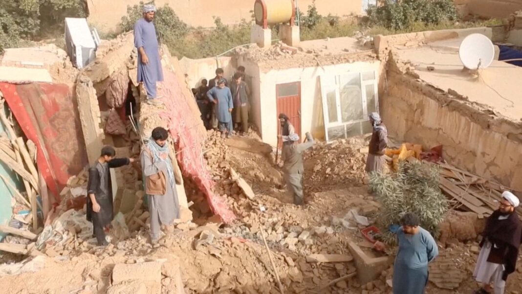A fotografia mostra uma cena de destruição massiva após um terremoto no Afeganistão, em uma área de clima seco. Há um grupo de cerca de dez homens no local, todos usando vestimentas tradicionais da região (túnicas longas, calças largas e, em alguns casos, turbantes ou lenços enrolados na cabeça). Eles parecem ser moradores ou voluntários, envolvidos em trabalhos de resgate ou busca.
