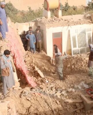 A fotografia mostra uma cena de destruição massiva após um terremoto no Afeganistão, em uma área de clima seco. Há um grupo de cerca de dez homens no local, todos usando vestimentas tradicionais da região (túnicas longas, calças largas e, em alguns casos, turbantes ou lenços enrolados na cabeça). Eles parecem ser moradores ou voluntários, envolvidos em trabalhos de resgate ou busca.