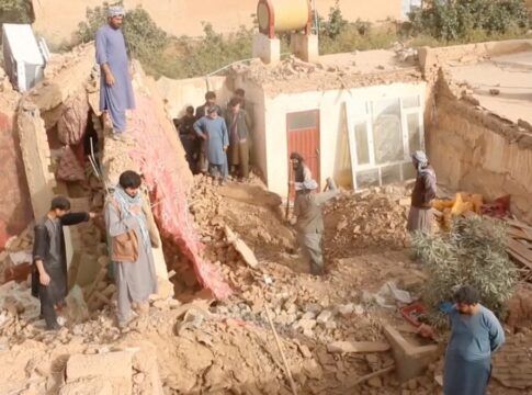 A fotografia mostra uma cena de destruição massiva após um terremoto no Afeganistão, em uma área de clima seco. Há um grupo de cerca de dez homens no local, todos usando vestimentas tradicionais da região (túnicas longas, calças largas e, em alguns casos, turbantes ou lenços enrolados na cabeça). Eles parecem ser moradores ou voluntários, envolvidos em trabalhos de resgate ou busca.