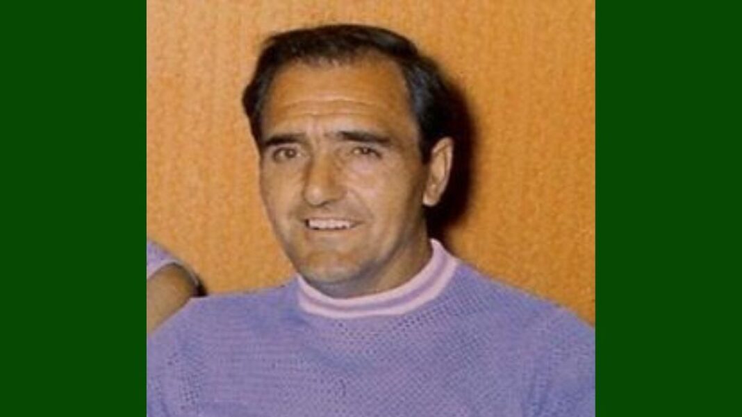 A foto é um retrato em close-up de um homem de meia-idade identificado como Alex Mitchell, da década de 1970. O homem tem cabelo escuro, curto e penteado de forma clássica. Ele parece ter uma tez clara e um sorriso aberto, que revela os dentes. As sobrancelhas são grossas e escuras.