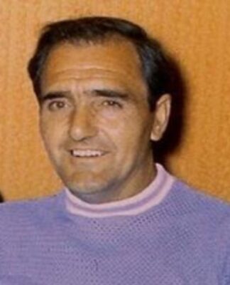 A foto é um retrato em close-up de um homem de meia-idade identificado como Alex Mitchell, da década de 1970. O homem tem cabelo escuro, curto e penteado de forma clássica. Ele parece ter uma tez clara e um sorriso aberto, que revela os dentes. As sobrancelhas são grossas e escuras.