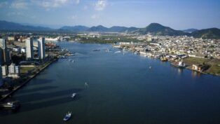 Esta é uma fotografia panorâmica, tirada de um ponto alto sob forte luz do dia, mostrando uma paisagem urbana, portuária e natural, dividindo as cidades de Santos e Guarujá.