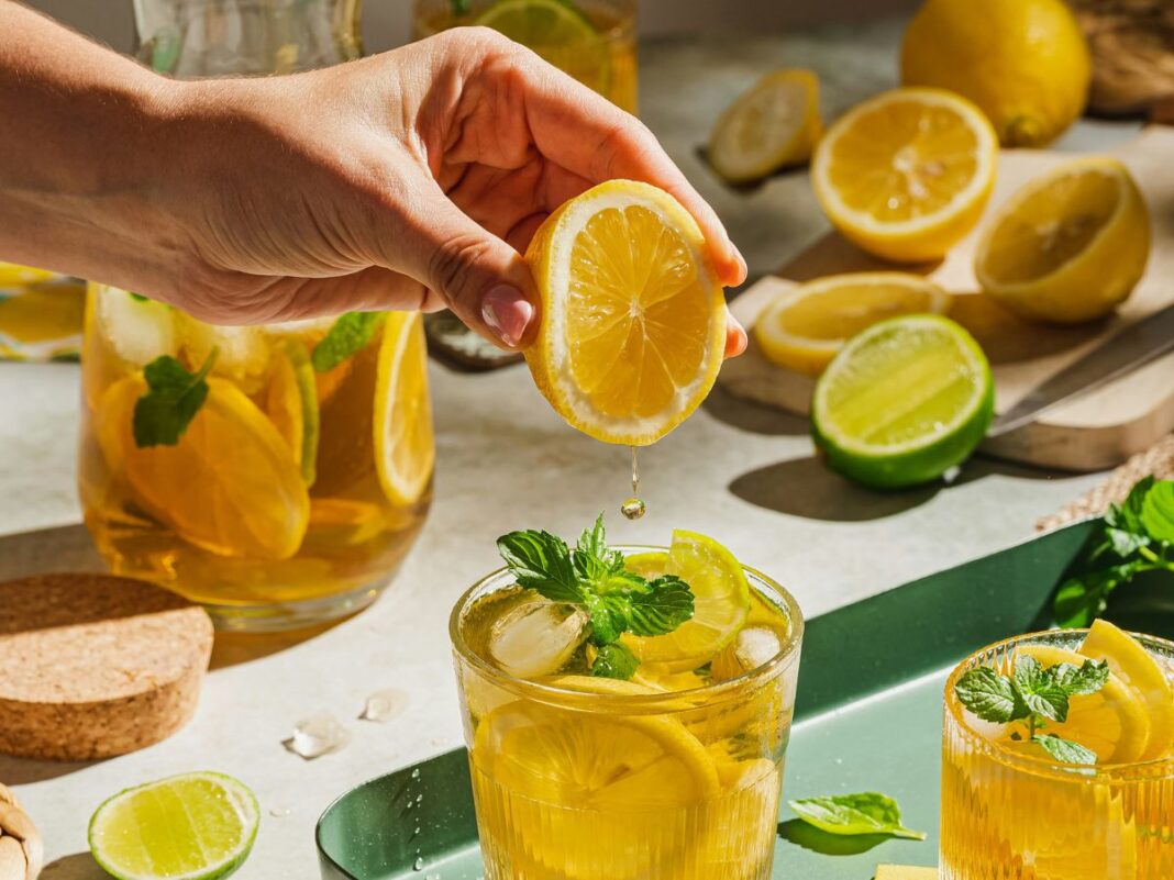 A imagem mostra uma pessoa espremendo uma laranja em um copo com chá que está em cima de uma bandeja verde