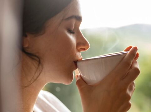 Na foto há uma mulher segurando e levando para a boca uma xícara branca.
