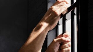 Imagem mostra as mãos de um detento segurando as grades da prisão