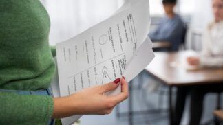 Em imagem ilustrativa, professora verifica prova, enquanto outro alunos estão realizando a mesma.