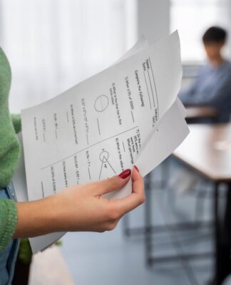 Em imagem ilustrativa, professora verifica prova, enquanto outro alunos estão realizando a mesma.