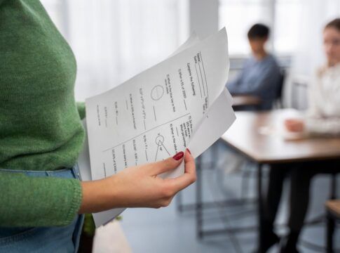 Em imagem ilustrativa, professora verifica prova, enquanto outro alunos estão realizando a mesma.