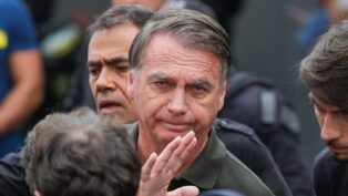 Imagem mostra o ex-presidente Jair Bolsonaro em primeiro plano. Atrás dele, aparece metade do rosto de um homem.