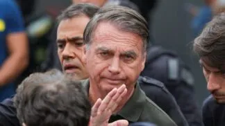 Imagem mostra o ex-presidente Jair Bolsonaro em primeiro plano. Atrás dele, aparece metade do rosto de um homem.