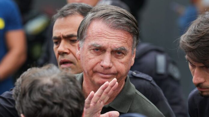Imagem mostra o ex-presidente Jair Bolsonaro em primeiro plano. Atrás dele, aparece metade do rosto de um homem.