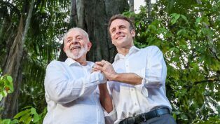 A imagem, com uma iluminação natural e suave, apresenta o Presidente do Brasil, Luiz Inácio Lula da Silva, e o Presidente da França, Emmanuel Macron, de pé, lado a lado, em um ambiente que parece ser uma floresta, possivelmente a Amazônia, como indicam os grandes troncos de árvores e a folhagem densa e escura no fundo.