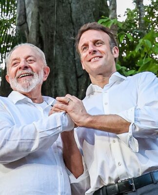 A imagem, com uma iluminação natural e suave, apresenta o Presidente do Brasil, Luiz Inácio Lula da Silva, e o Presidente da França, Emmanuel Macron, de pé, lado a lado, em um ambiente que parece ser uma floresta, possivelmente a Amazônia, como indicam os grandes troncos de árvores e a folhagem densa e escura no fundo.